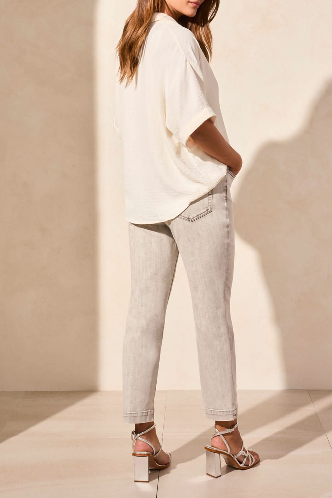 A woman stands indoors, facing away, wearing the Tribal FLOWY POPOVER SHORT SLEEVE SHIRT in white, light gray jeans, and metallic block-heeled sandals.