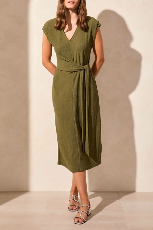 A woman models the Tribal SHORT SLEEVE DRESS WITH FRONT TIE in olive green, featuring a flattering silhouette and waist tie, standing against a neutral background with her hands behind her back.