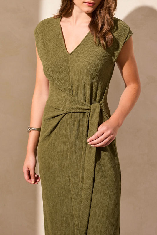 A woman models the Tribal SHORT SLEEVE DRESS WITH FRONT TIE in olive green, featuring a flattering silhouette and waist tie, standing against a neutral background with her hands behind her back.