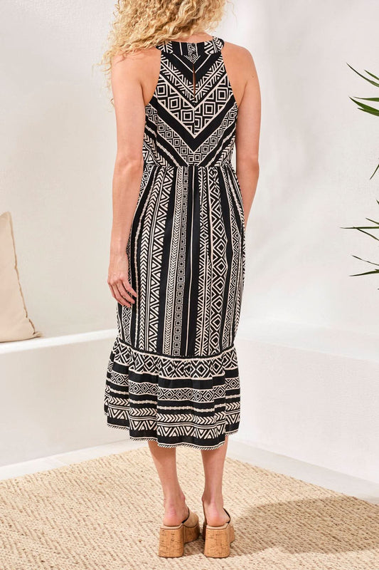 A woman stands against a stone wall, wearing the Tribal HALTER DRESS—sleeveless with a black and white pattern—and black sandals, her hair down, one hand in her pocket.