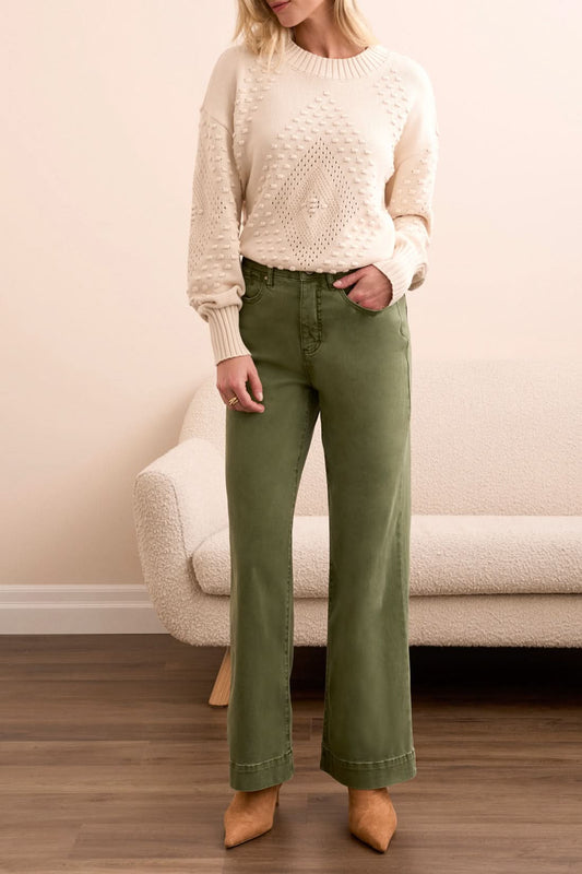 A woman stands indoors in Tribal FLY FRONT WIDE LEG PANTS, a cream knit sweater, and tan heels, posed by a light sofa on a wooden floor.