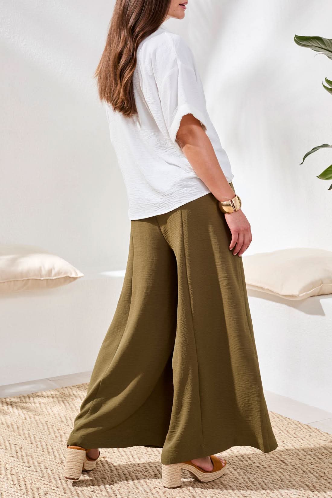 Indoors, a woman wears Tribal Wide Leg Flowy Pant in olive green, paired with a loose white short-sleeve blouse, tan sandals, and a gold bracelet.