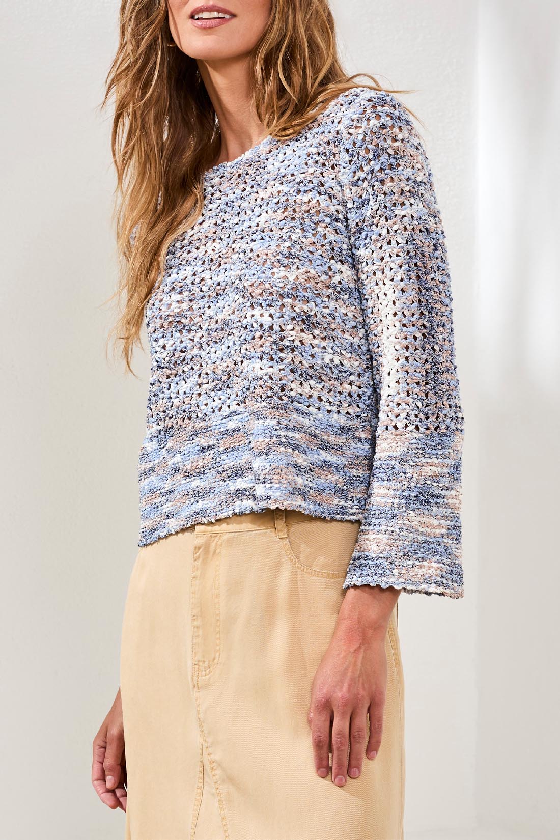 A woman wears the Tribal Bell Sleeve Sweater—an open stitch, long-sleeve blue and beige top—paired with a high-waisted beige skirt, shown from shoulders to knees.