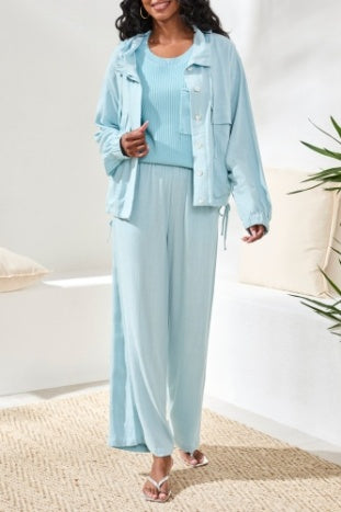 A woman wears the Tribal Light Jacket with Hood, a light blue button-up style paired with matching pants, ideal for effortless layering near green plants in a bright indoor space.