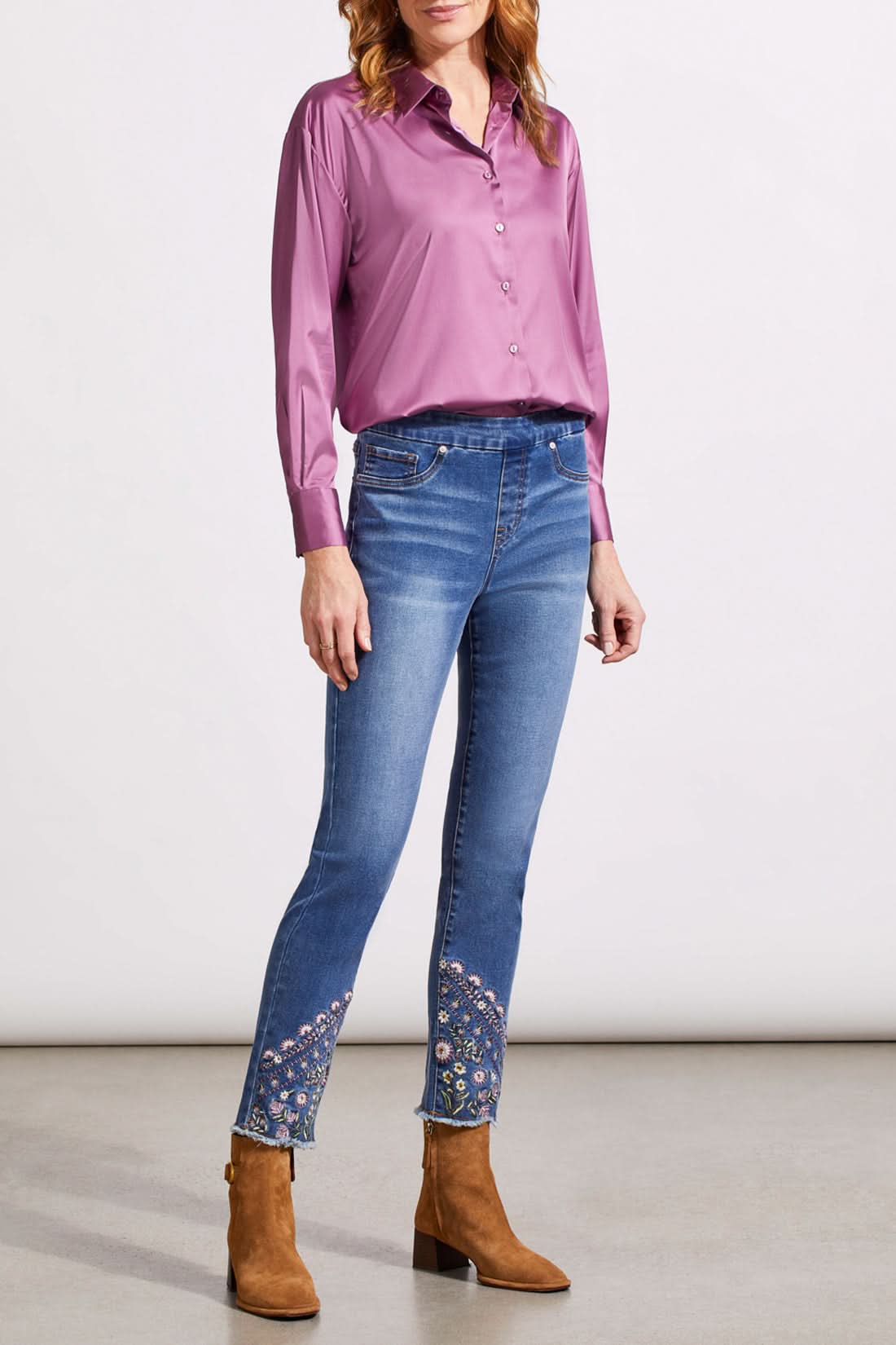 A person stands wearing a long-sleeve pink blouse, Tribal's Audrey Pull on Slim Ankle with Fancy Embroidery blue jeans, and slim ankle brown boots. The background is a plain, light-colored wall and floor.