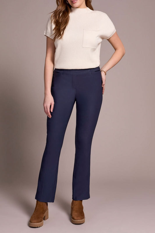 A woman in a beige turtleneck, Tribal FLATTEN IT PULL ON PANT, and brown boots stands before a cream sofa on a wooden floor.