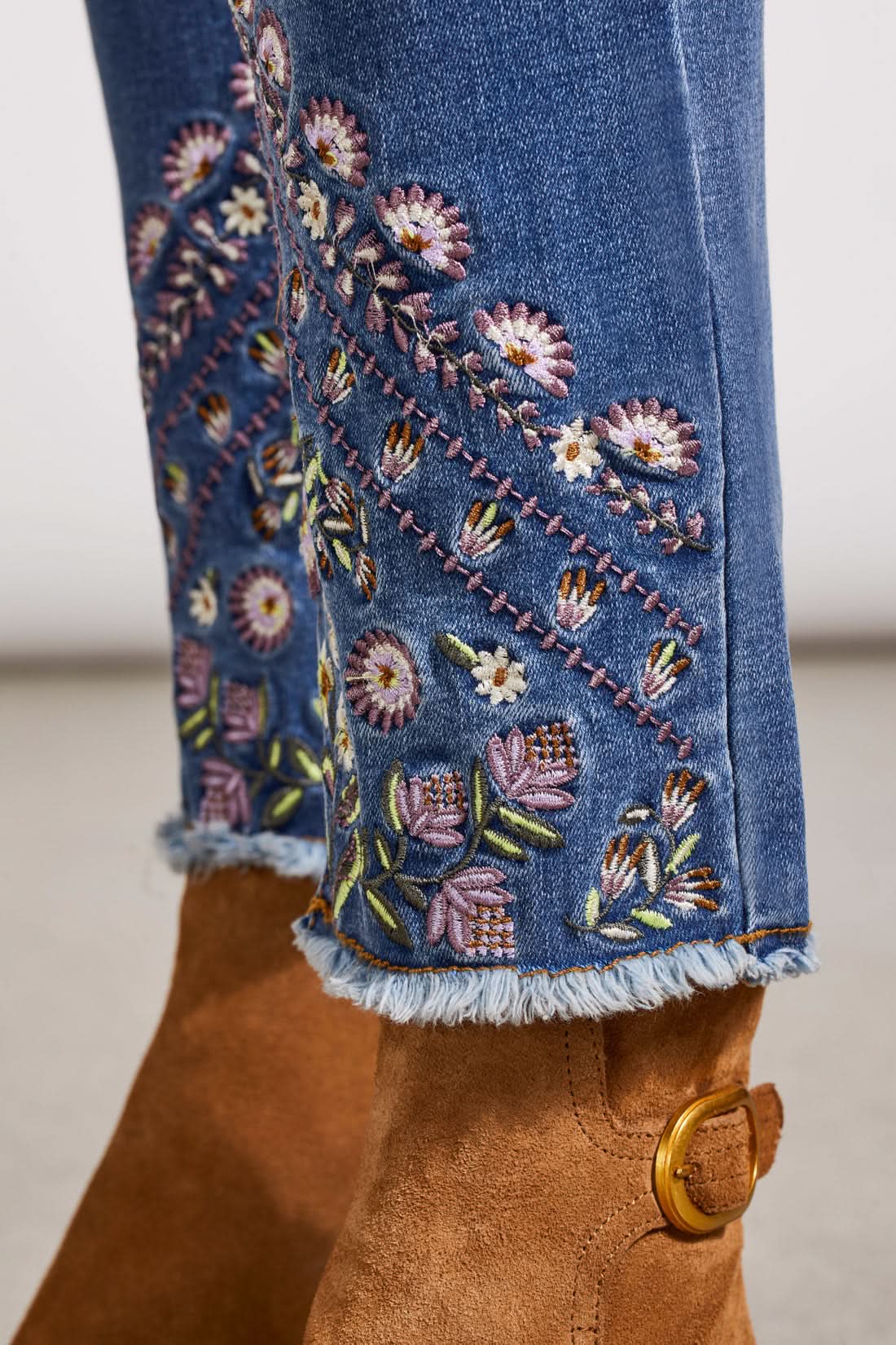 A person stands wearing a long-sleeve pink blouse, Tribal's Audrey Pull on Slim Ankle with Fancy Embroidery blue jeans, and slim ankle brown boots. The background is a plain, light-colored wall and floor.