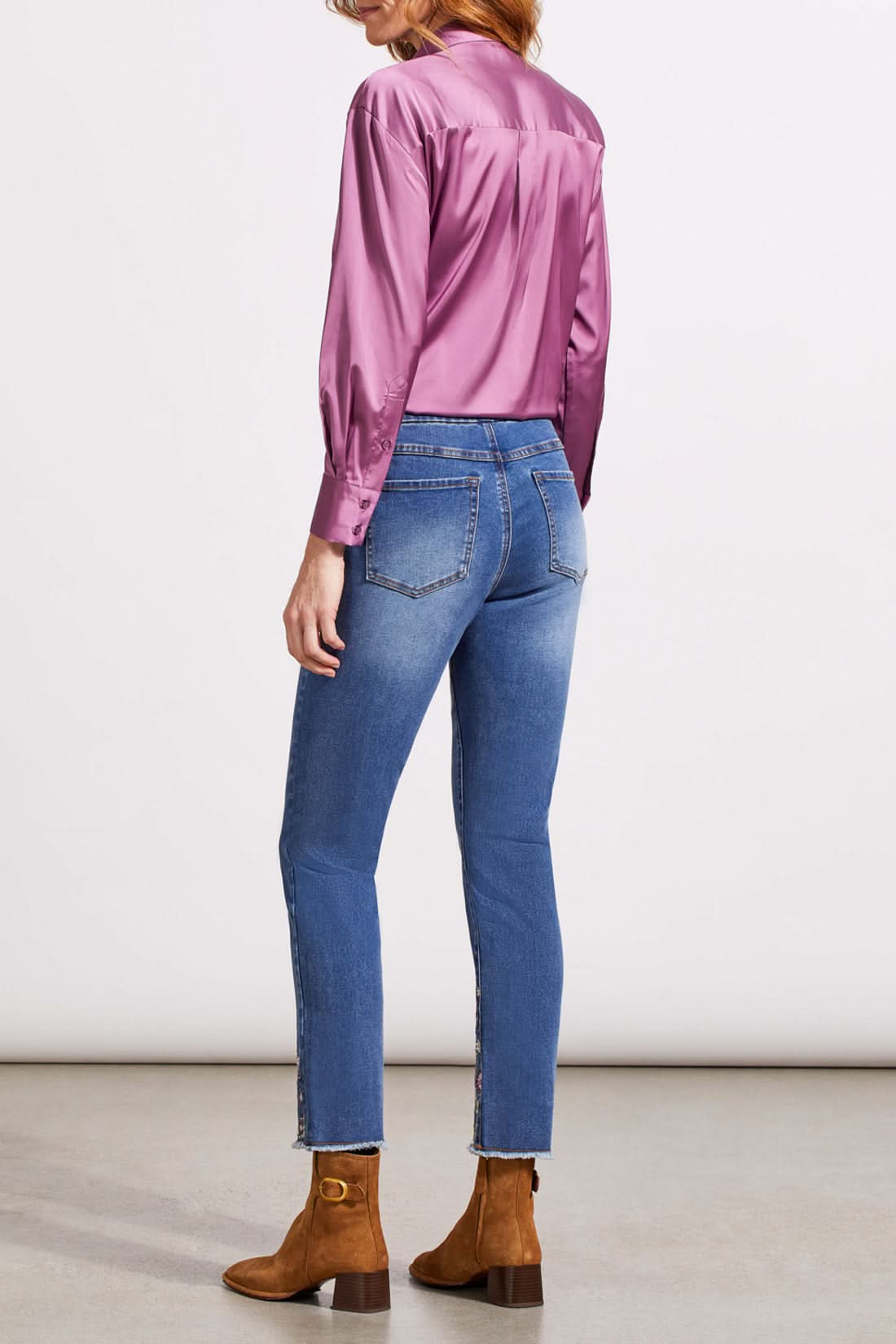 A person stands wearing a long-sleeve pink blouse, Tribal's Audrey Pull on Slim Ankle with Fancy Embroidery blue jeans, and slim ankle brown boots. The background is a plain, light-colored wall and floor.