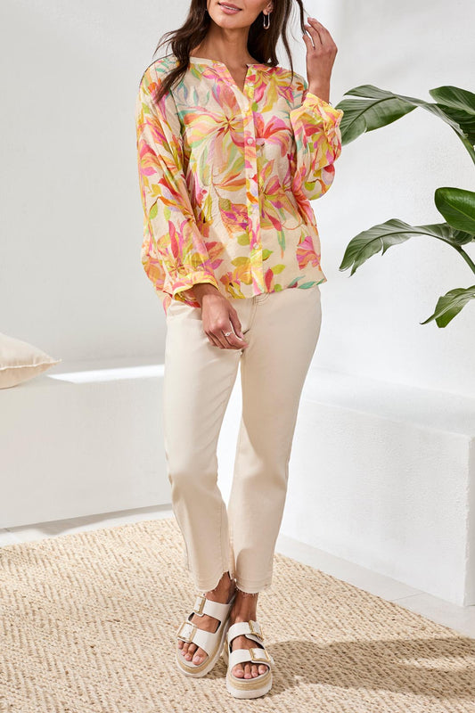 A woman with long blonde hair wears the Tribal Breezy Summer Blouse while sitting outdoors and smiling. Colorful cushions and greenery can be seen in the background.