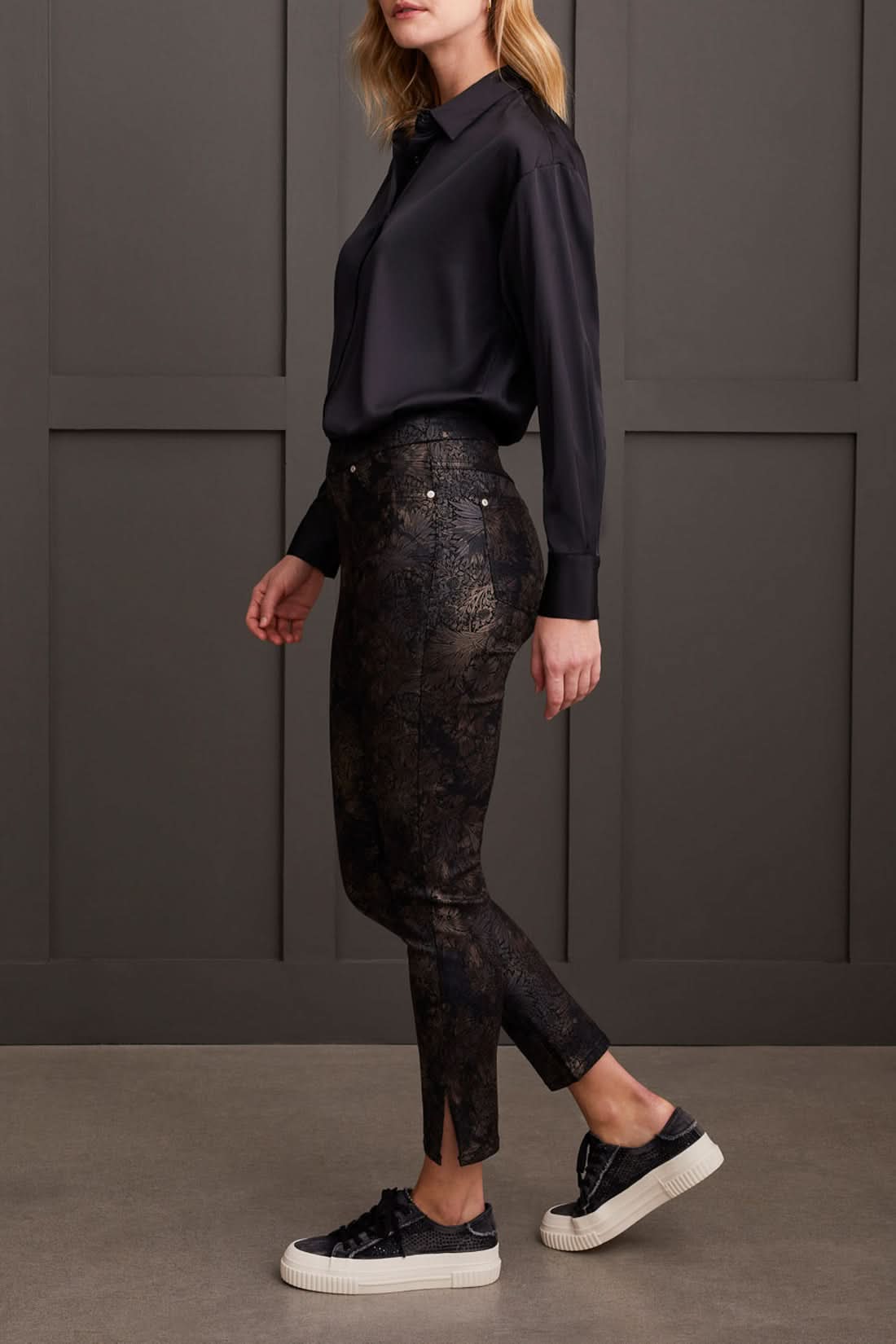 Against a paneled wall, a person stands sideways wearing a black blouse, Tribal Audrey Pull on Slim Ankle pants with a side slit in black patterned design, and black platform sneakers.