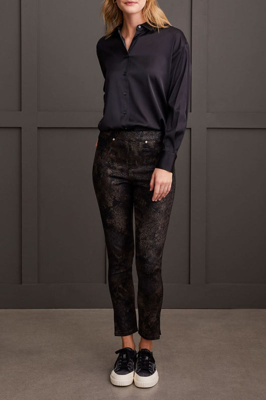 Against a paneled wall, a person stands sideways wearing a black blouse, Tribal Audrey Pull on Slim Ankle pants with a side slit in black patterned design, and black platform sneakers.