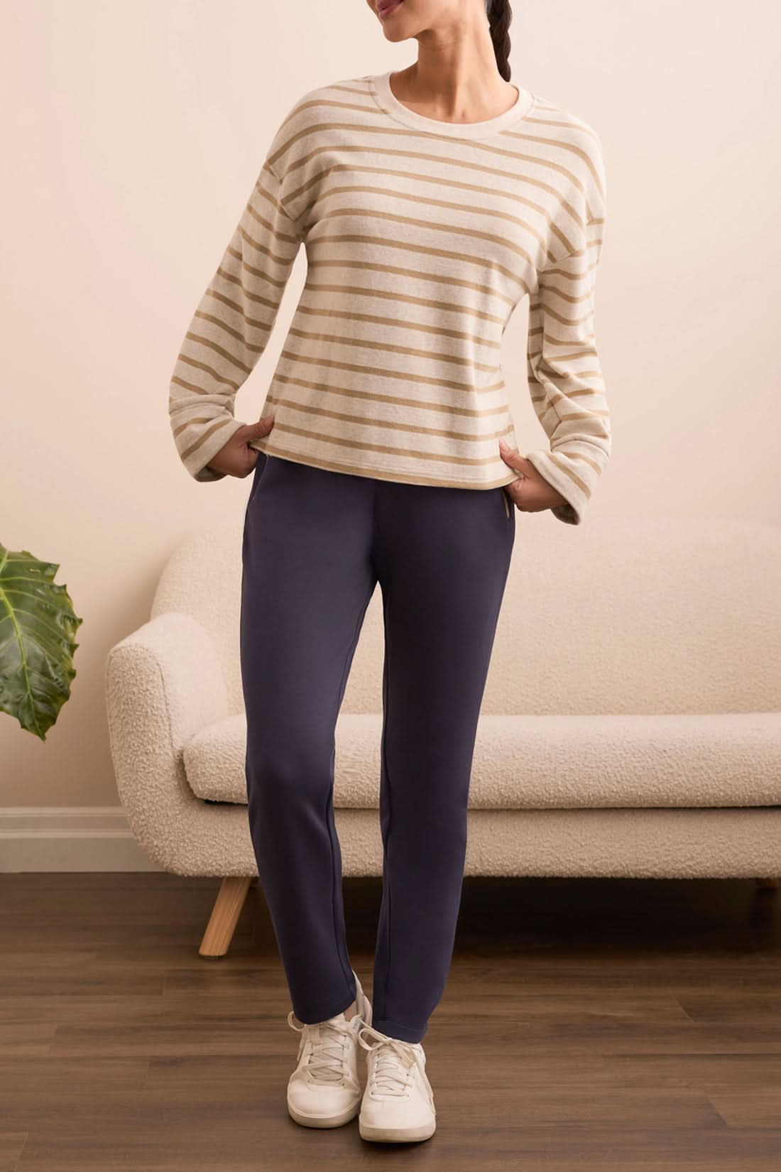 Indoors, a person stands near a potted plant and cream sofa wearing a beige striped long-sleeve shirt, white sneakers, and Tribal CONTRAST TRIM PULL-ON ANKLE PANTS with one hand in their pocket.
