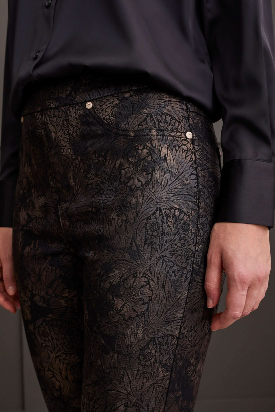 Against a paneled wall, a person stands sideways wearing a black blouse, Tribal Audrey Pull on Slim Ankle pants with a side slit in black patterned design, and black platform sneakers.