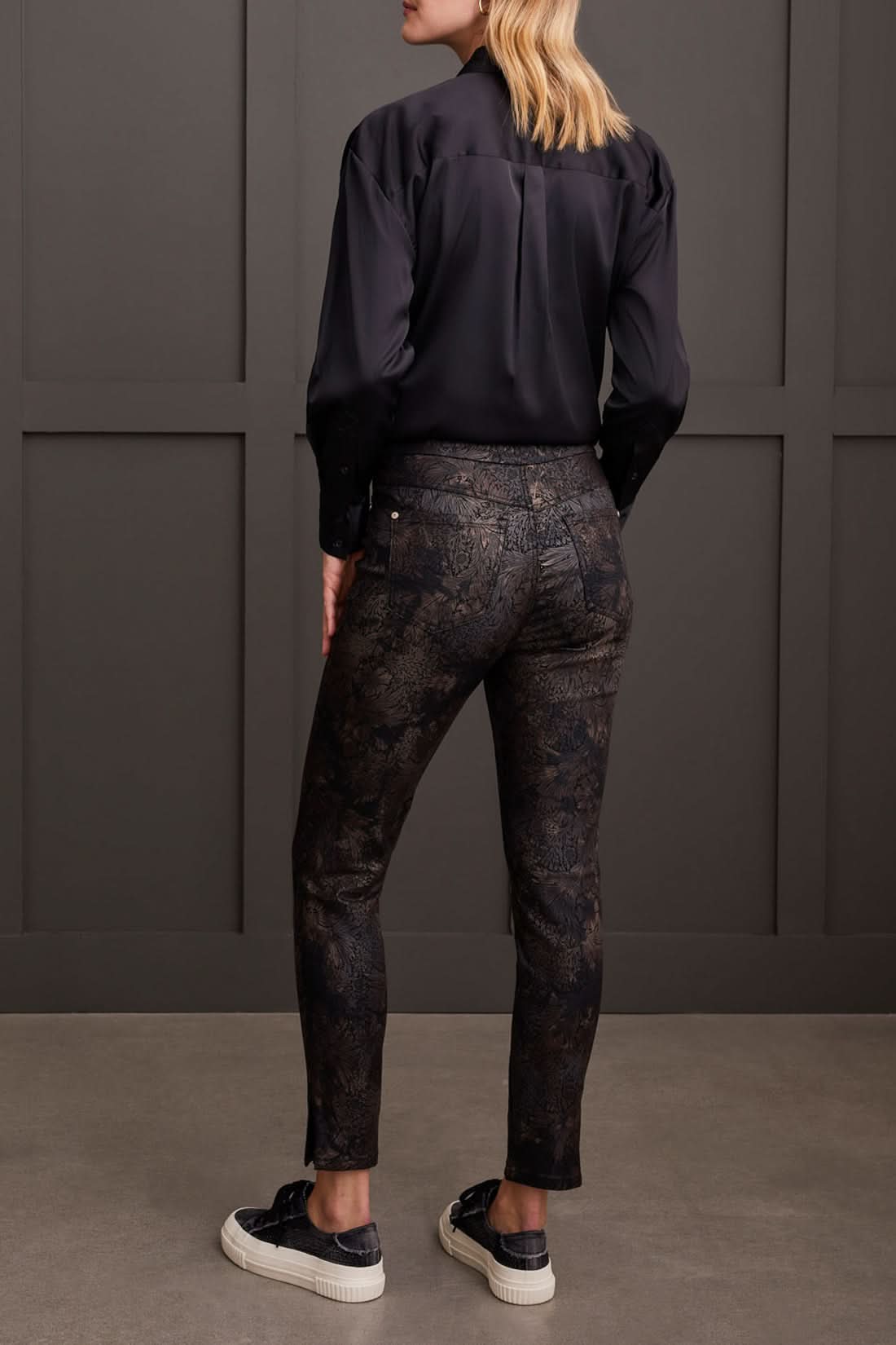Against a paneled wall, a person stands sideways wearing a black blouse, Tribal Audrey Pull on Slim Ankle pants with a side slit in black patterned design, and black platform sneakers.