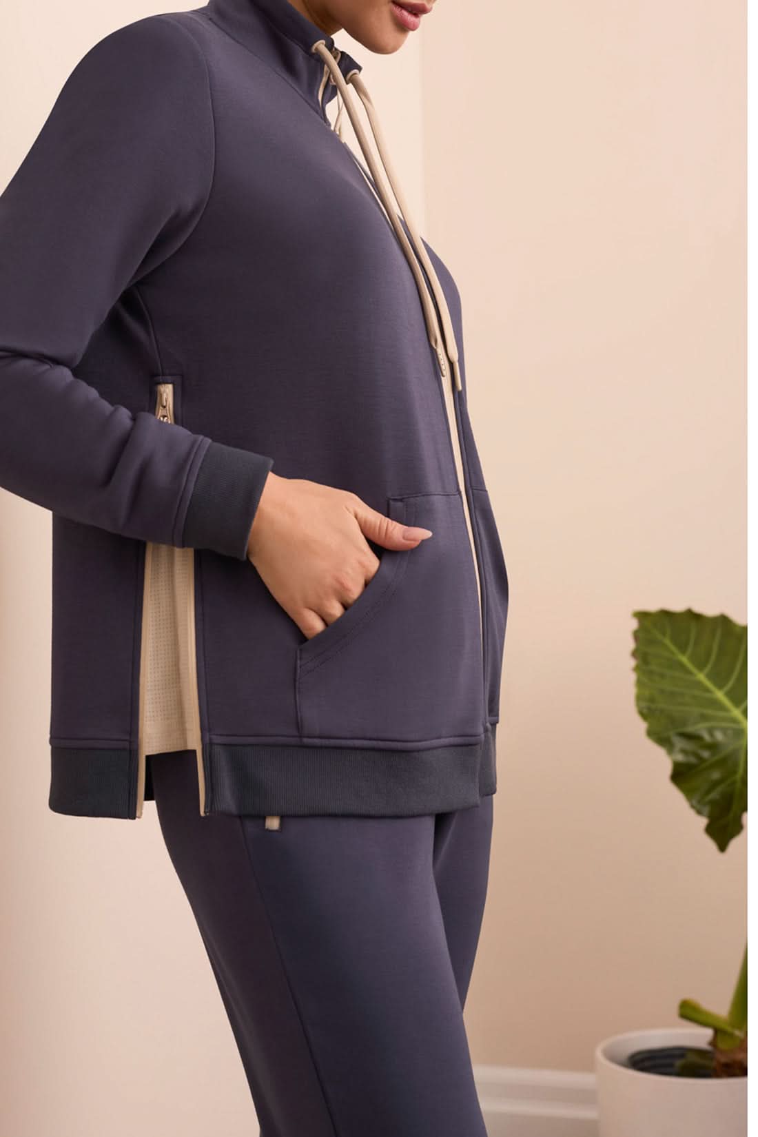 A person stands indoors in a navy tracksuit paired with the Tribal MOCK NECK CARDIGAN in beige and white stripes and white sneakers, with a green plant and cream-colored sofa in the background.