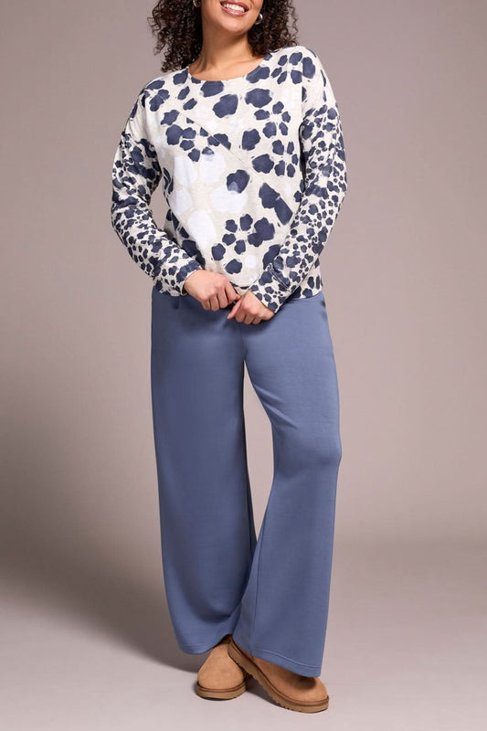 Someone stands against a neutral background wearing the Tribal CREW NECK TOP with a white and navy animal print, wide-leg blue pants, and tan slippers—an ideal look for casual wear.