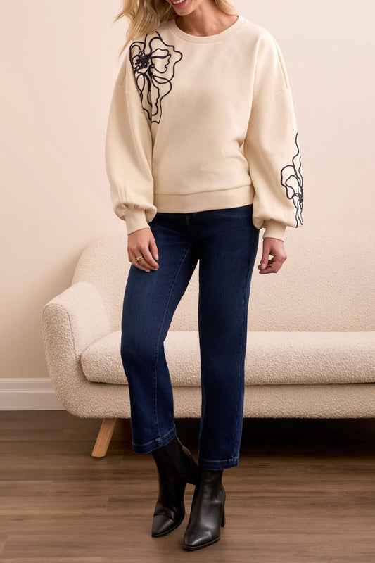 Indoors, a woman wears Tribal’s CREW NECK TOP W/EMBROIDERY—cream with black floral details and balloon sleeves—paired with dark blue jeans and black ankle boots beside a beige textured sofa.