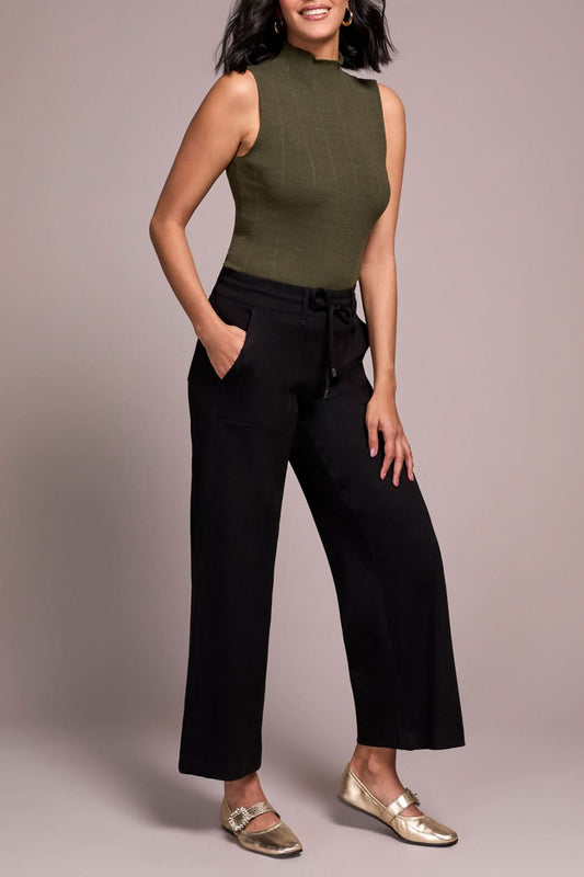 A person wearing a sleeveless olive green mock neck top, Tribal Dark Ivy Drawcord Pant in black, and gold loafers stands against a plain background.