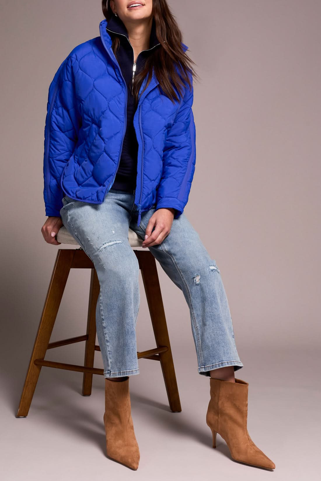 A person in the Tribal ZIPPED FRT QUILTED JACKET in lapis blue, a navy sweater, light blue distressed jeans, and tan heeled boots stands against a plain background.