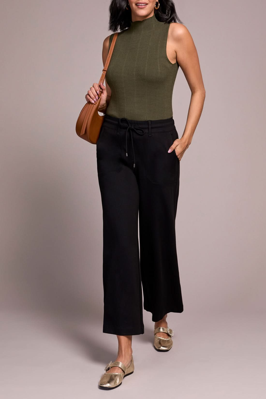 A person wearing a sleeveless olive green mock neck top, Tribal Dark Ivy Drawcord Pant in black, and gold loafers stands against a plain background.