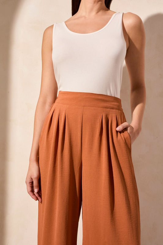 A woman stands against a neutral background wearing a rust-colored button-up shirt, a white top, brown heeled sandals, and Tribal’s FLOWY PANT WITH BLACK ELASTIC WAISTBAND.