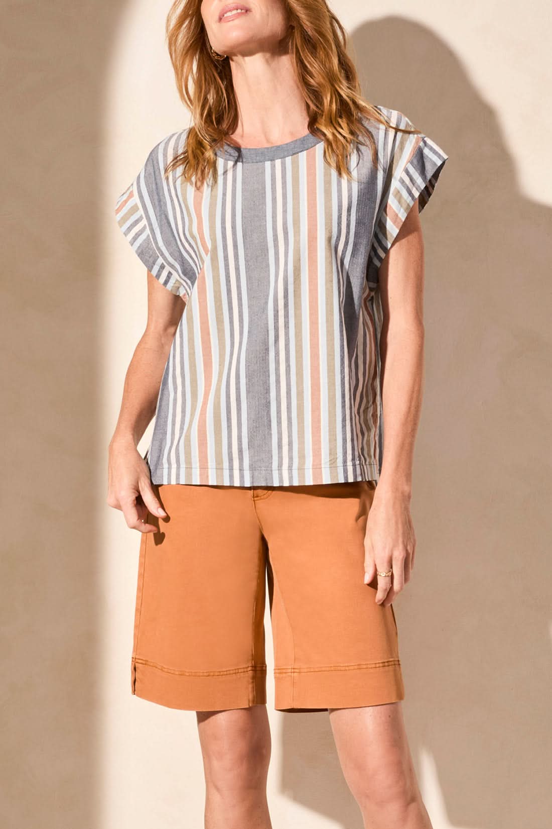 A woman stands against a neutral wall wearing Tribal's DROP SHOULDER GUSSET SLEEVE TOP with a flattering fit, paired with knee-length orange shorts.