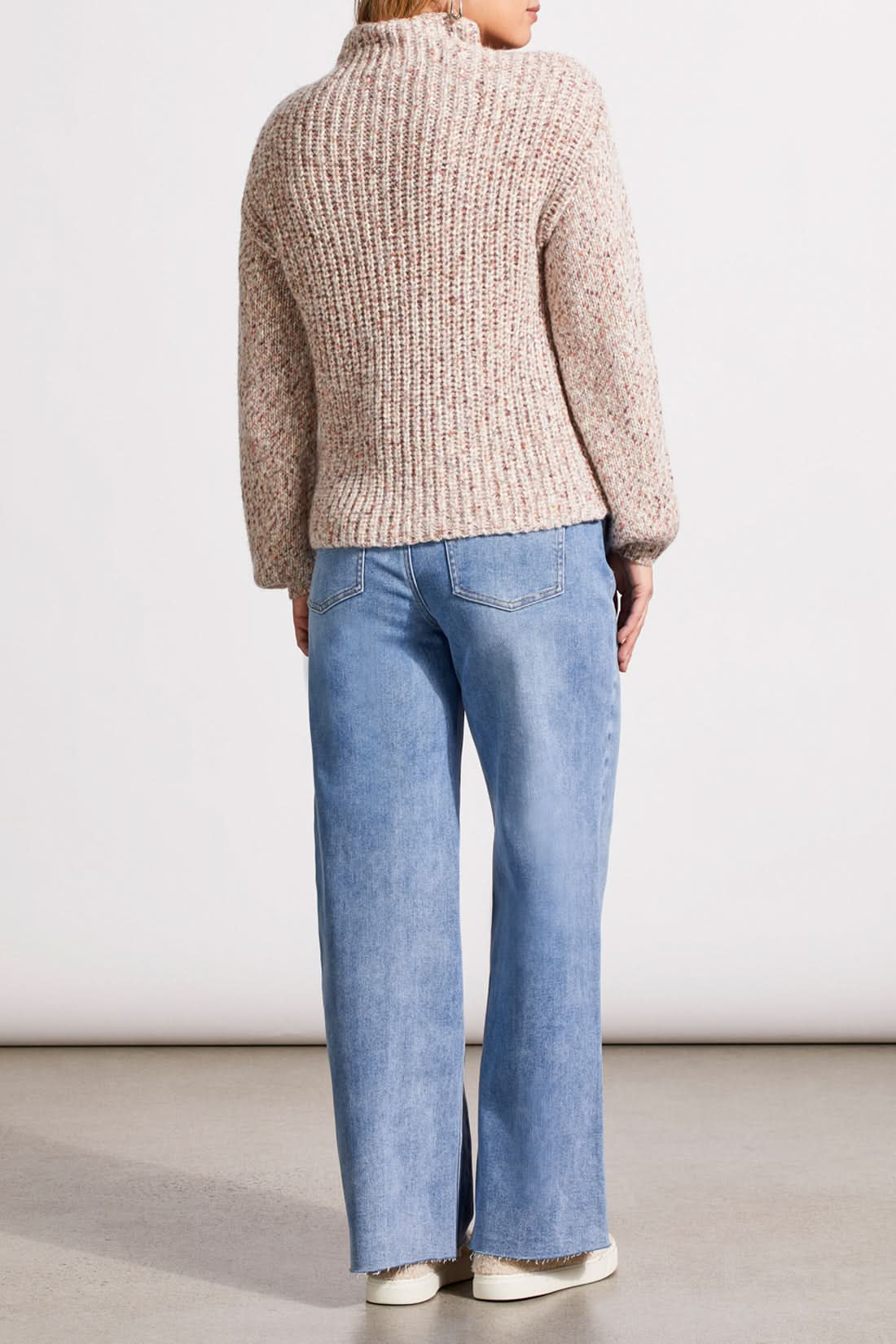 Person wearing a red patterned coat, cozy Tribal Funnel Neck Oversize Sweater in red, blue jeans, and black ankle boots stands against a dark paneled wall.