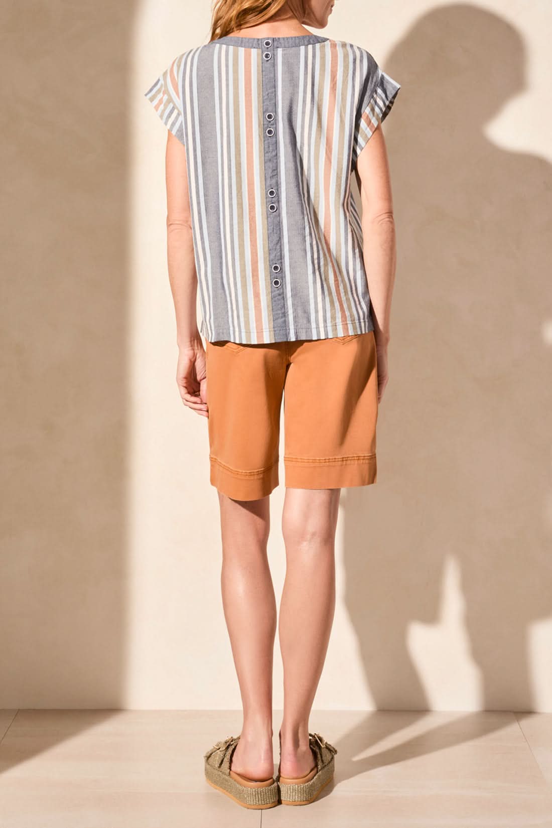 A woman stands against a neutral wall wearing Tribal's DROP SHOULDER GUSSET SLEEVE TOP with a flattering fit, paired with knee-length orange shorts.
