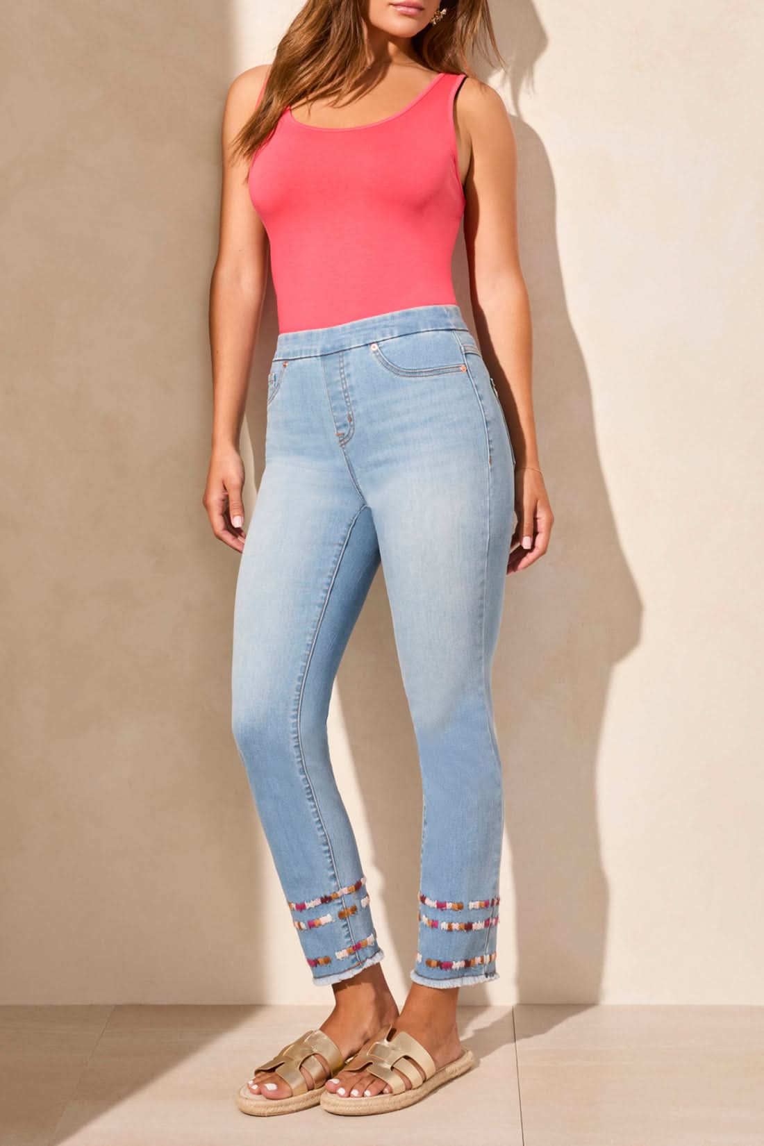 A woman stands against a beige wall wearing a sleeveless coral top, Tribal AUDREY PULL ON SLIM ANKLE JEANS with beaded hems for a flattering fit, and beige sandals—blending comfort and effortless style.