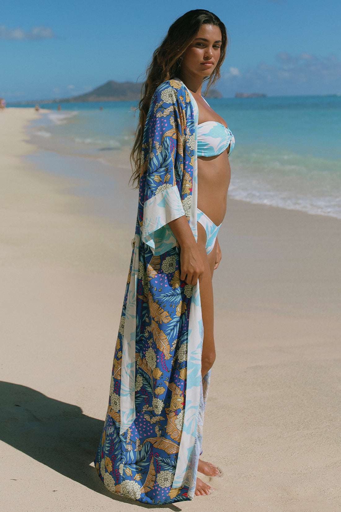 A woman in a blue and white bikini and the Tribal 3/4 SLEEVE DUSTER COVER UP strolls along a sandy beach, exuding chic vacation style with the ocean and a distant island behind her.