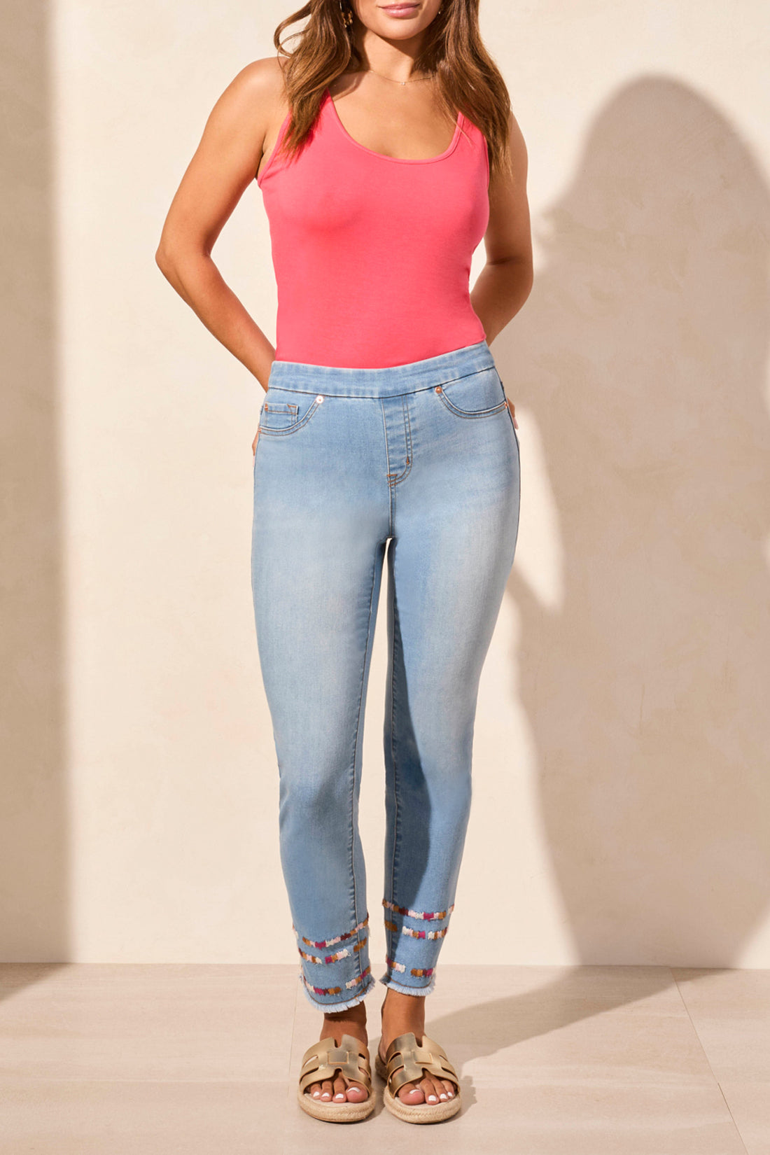 A woman stands against a beige wall wearing a sleeveless coral top, Tribal AUDREY PULL ON SLIM ANKLE JEANS with beaded hems for a flattering fit, and beige sandals—blending comfort and effortless style.