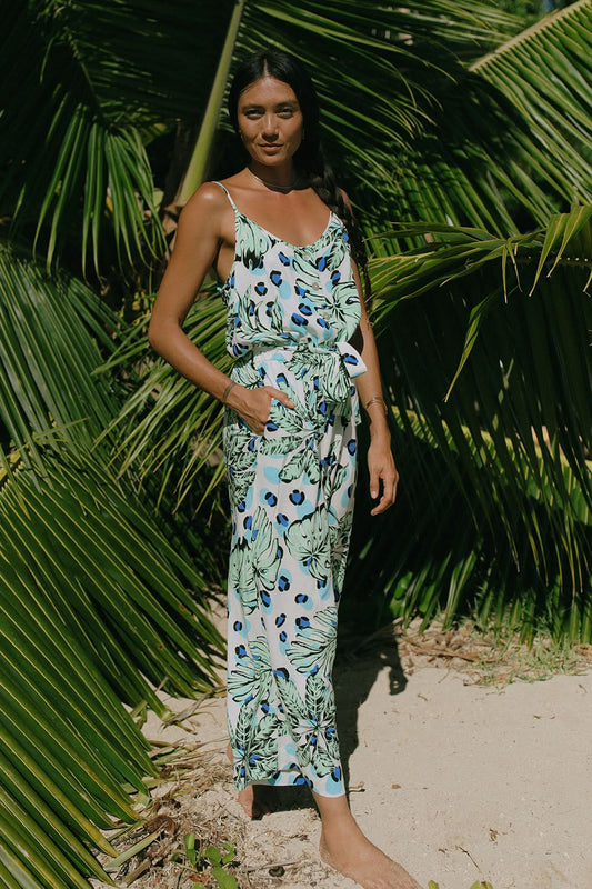 A woman stands barefoot on sand, wearing the Tribal BUTTON FRONT JUMPSUIT WITH POCKETS in a sleeveless tropical print—an effortless summer style surrounded by lush green palm leaves.