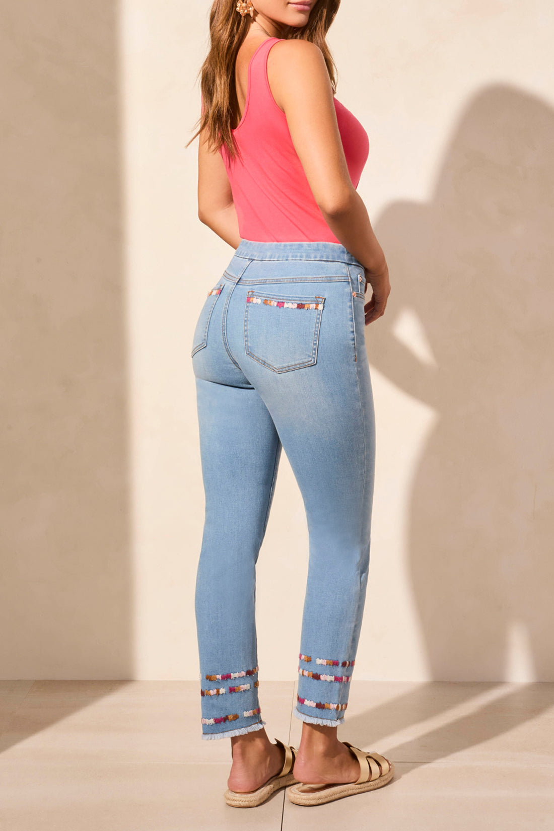 A woman stands against a beige wall wearing a sleeveless coral top, Tribal AUDREY PULL ON SLIM ANKLE JEANS with beaded hems for a flattering fit, and beige sandals—blending comfort and effortless style.