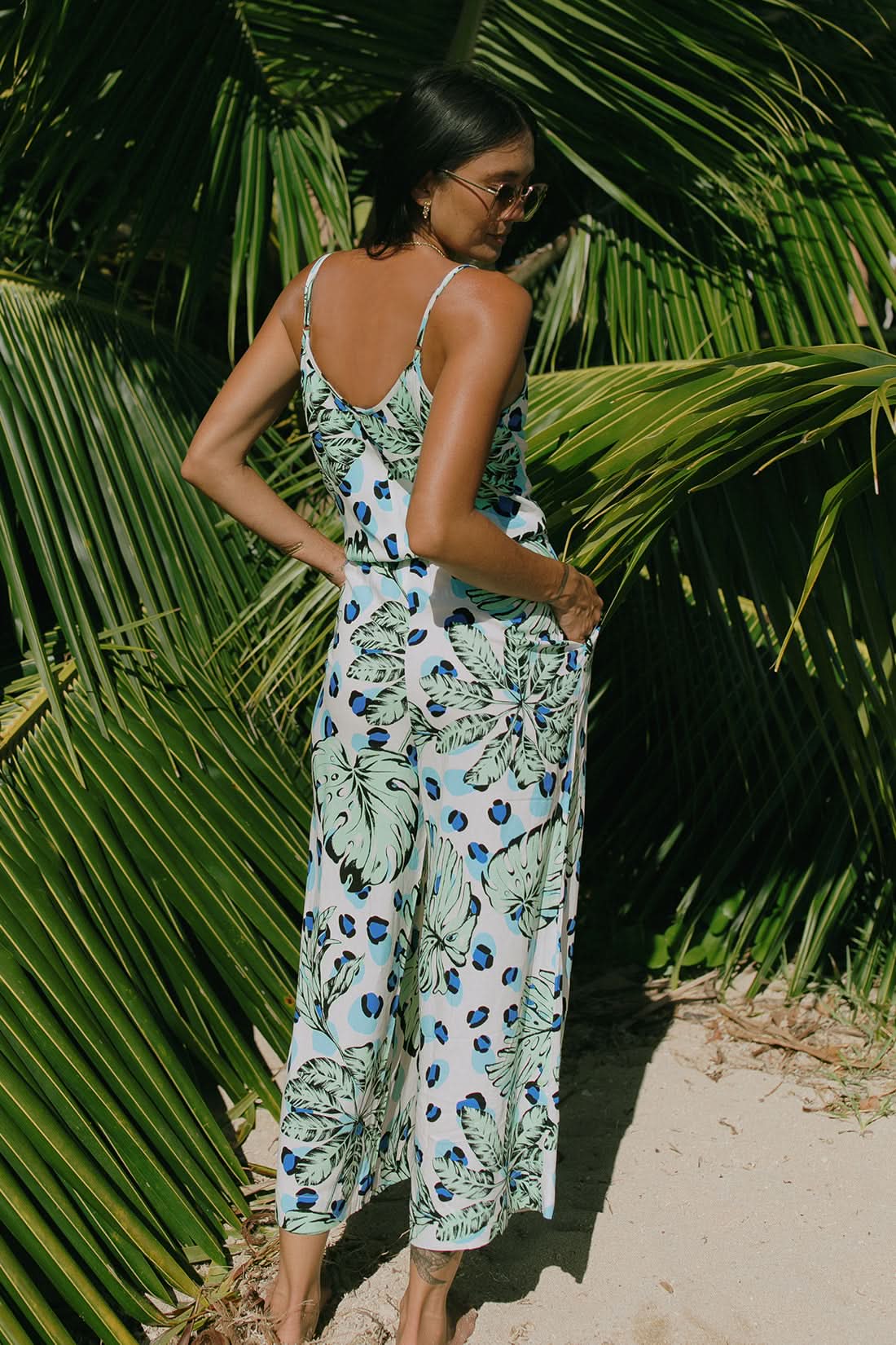 A woman stands barefoot on sand, wearing the Tribal BUTTON FRONT JUMPSUIT WITH POCKETS in a sleeveless tropical print—an effortless summer style surrounded by lush green palm leaves.