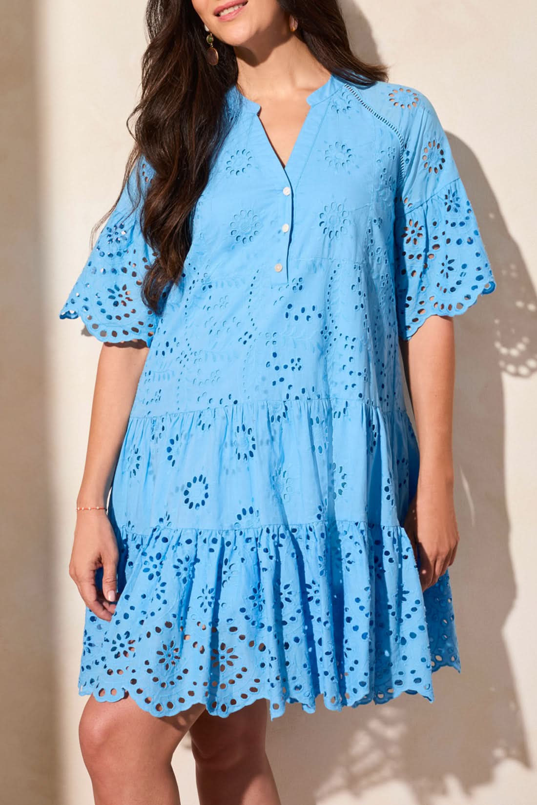 A woman stands against a light-colored wall, wearing the Tribal MIXED PATTERN EYELET DRESS—a light blue, short-sleeved, tiered summer dress with eyelet lace details, a V-neckline, and a scalloped hem.