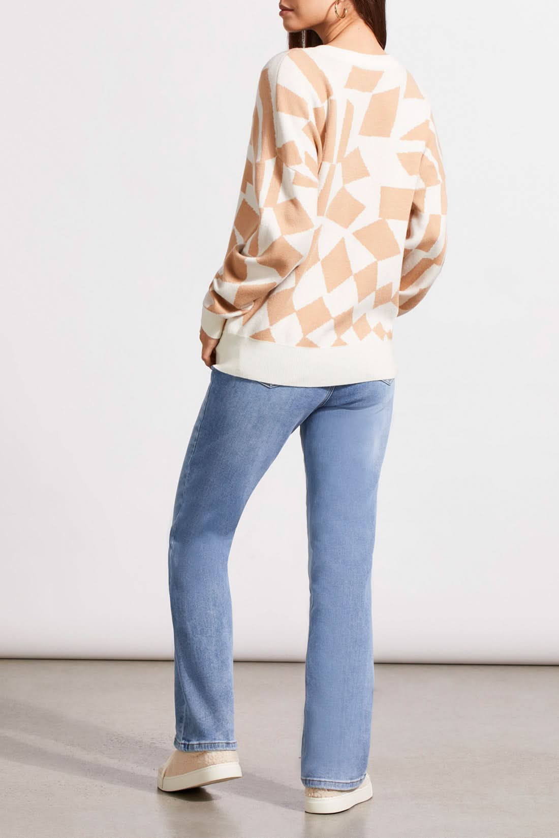 A person stands against a plain background wearing a cozy Tribal V-Neck Jacquard Sweater with a beige geometric pattern, light blue jeans, and beige sneakers. They have one hand in their pocket, effortlessly stylish.