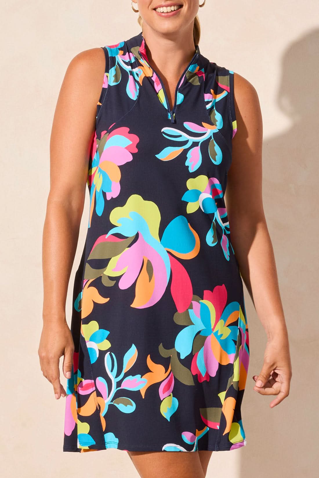 A woman stands against a beige wall wearing the Tribal SLEEVELESS DRESS WITH FLARE CUT, featuring a colorful floral pattern, paired with white socks and white sneakers.