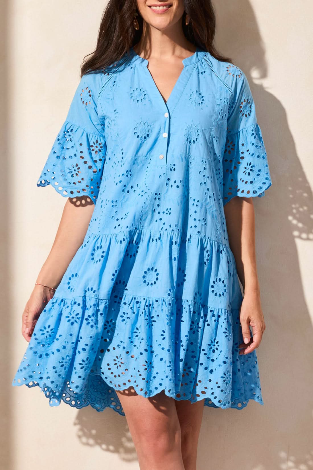 A woman stands against a light-colored wall, wearing the Tribal MIXED PATTERN EYELET DRESS—a light blue, short-sleeved, tiered summer dress with eyelet lace details, a V-neckline, and a scalloped hem.