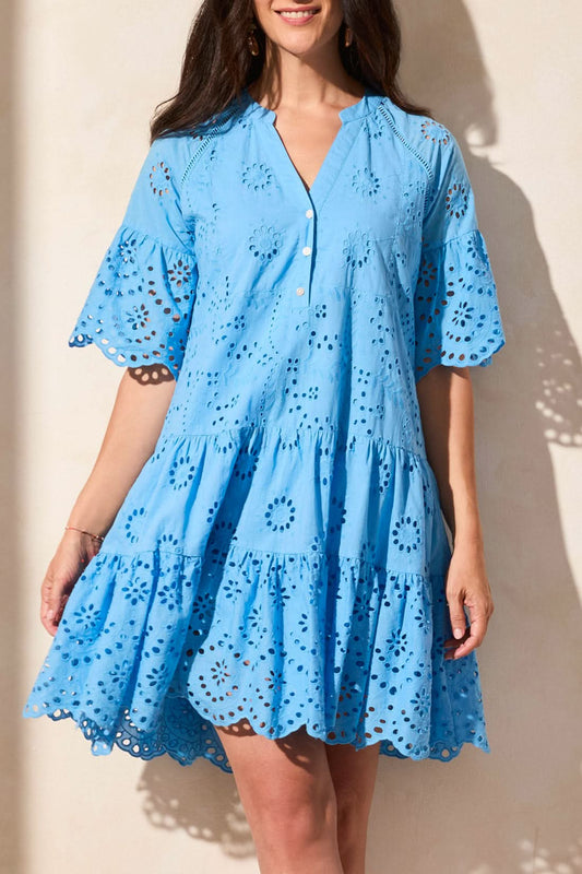 A woman stands against a light-colored wall, wearing the Tribal MIXED PATTERN EYELET DRESS—a light blue, short-sleeved, tiered summer dress with eyelet lace details, a V-neckline, and a scalloped hem.