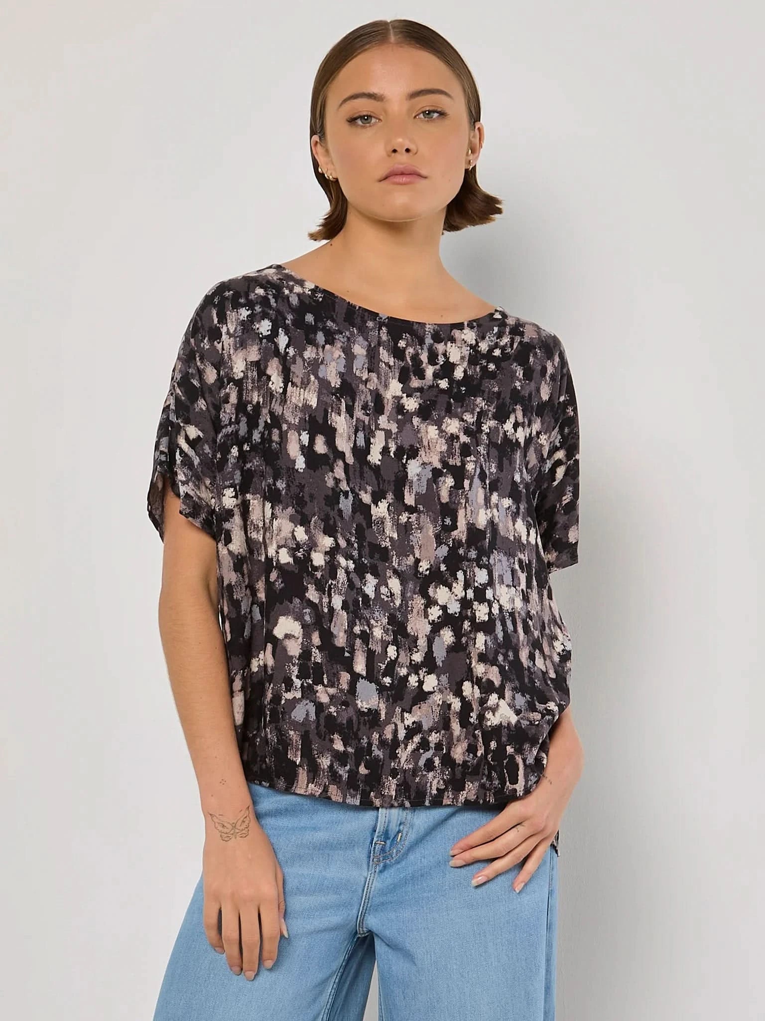 A person in an Apricot Batwing Brushstroke MK2 top and light jeans stands against a plain background.