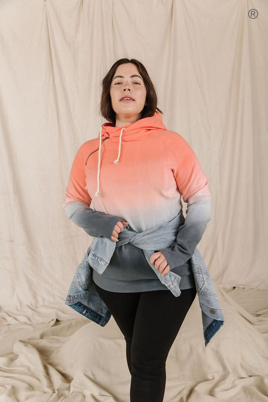 Dressed in Ampersand Avenue's Chasing Sunset Doublehooded Sweatshirt and denim shorts, a person exudes comfort and style while standing in a kitchen adorned with a marble backsplash and wooden cabinets.
