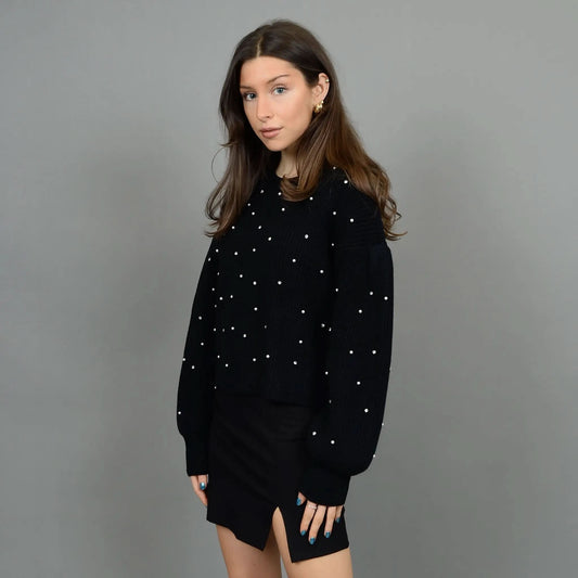 A person stands against a plain background wearing the stylish and cozy RD International Niri Long Sleeve Pull Over Sweater adorned with white dots, paired with a black skirt featuring a front slit. Their hands are in their pockets as they look slightly to their right.