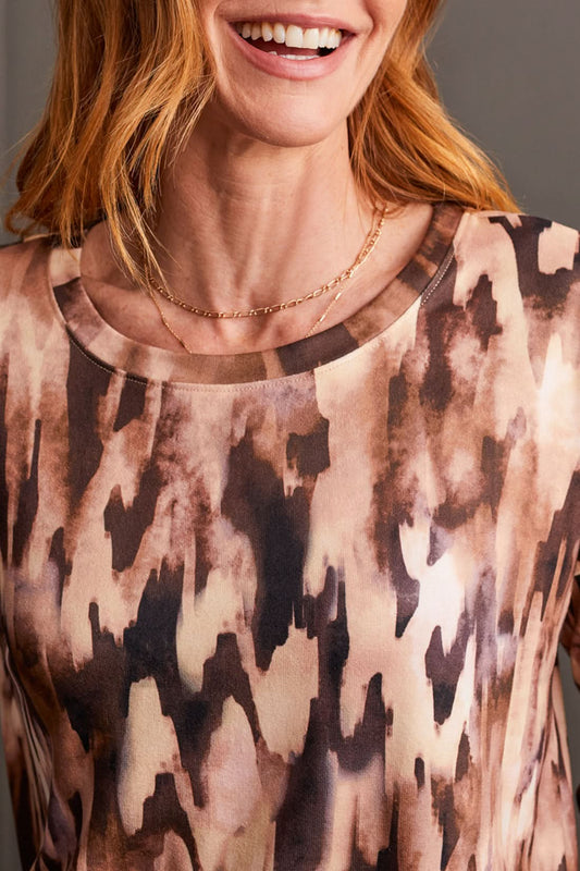 A person wearing the Tribal Crew Neck Top with Curved Hem, featuring a stylish patterned design, paired with a black knee-length skirt and black ankle boots, stands against a dark paneled wall—perfect for any fall wardrobe.