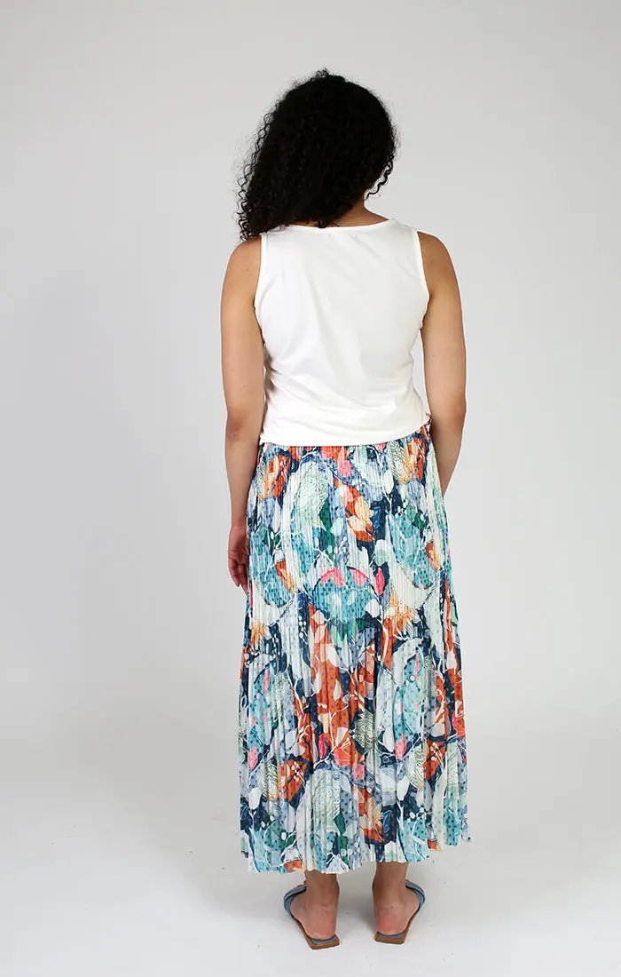 A woman in a white tank top and a playful Fresh FX pleated floral skirt smiles faintly while looking to the side.