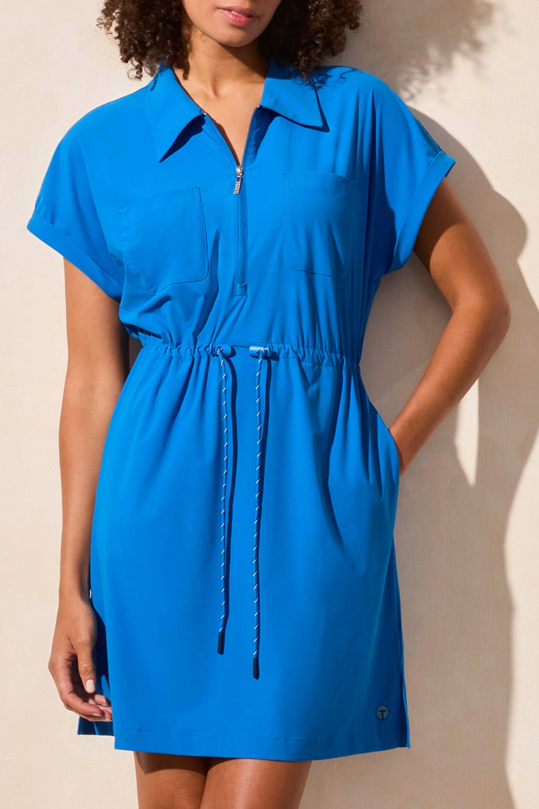 A person wears the Tribal SHORT SLEEVE ZIP POLO DRESS WITH DRAWCORD in bright blue, featuring a customizable fit and pockets, paired with white and blue sneakers, standing against a light-colored wall.