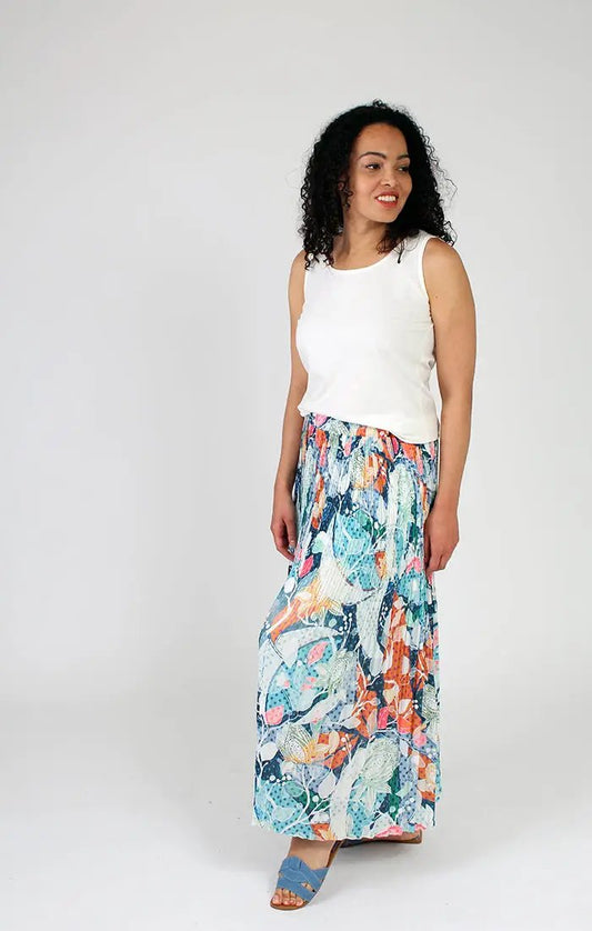 A woman in a white tank top and a playful Fresh FX pleated floral skirt smiles faintly while looking to the side.