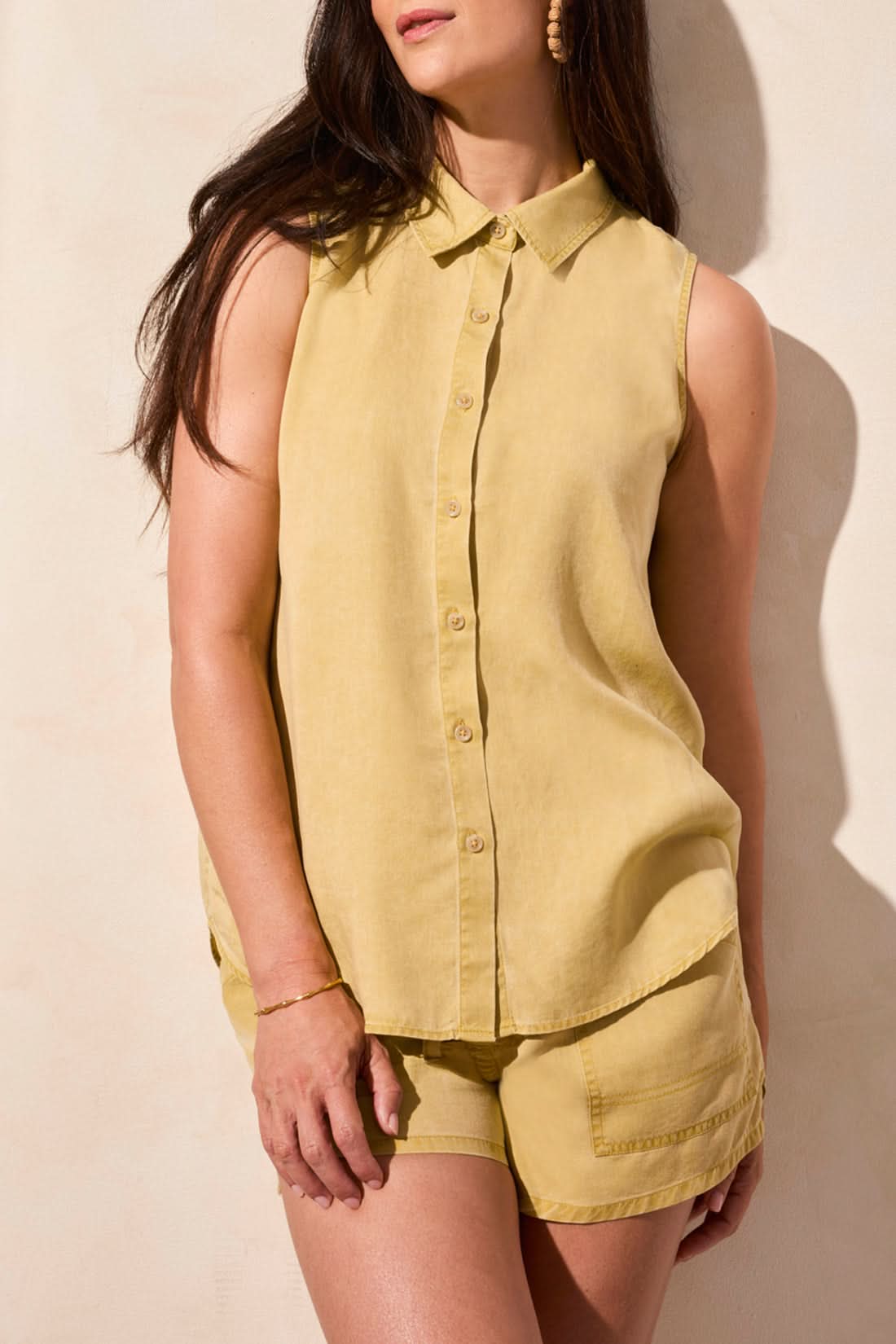 A woman stands against a light-colored wall wearing the Tribal FLOWY SLEEVELESS BUTTON DOWN SHIRT and matching yellow shorts.