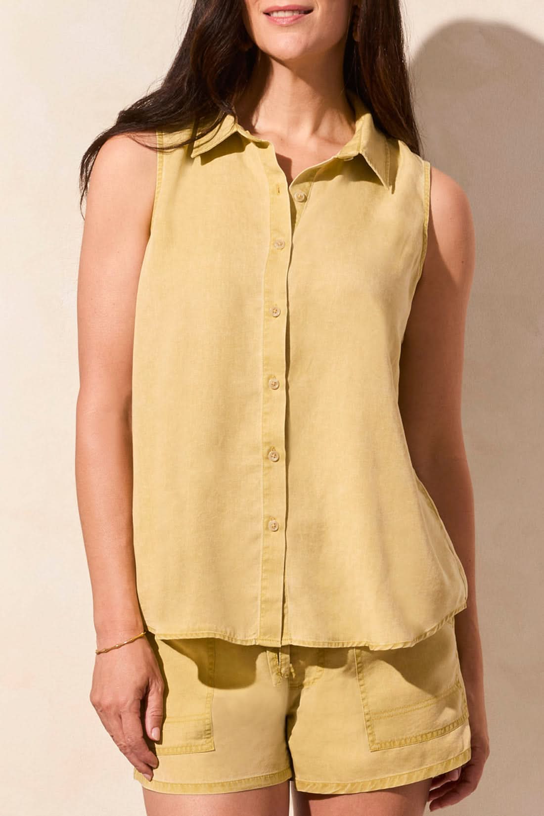 A woman stands against a light-colored wall wearing the Tribal FLOWY SLEEVELESS BUTTON DOWN SHIRT and matching yellow shorts.