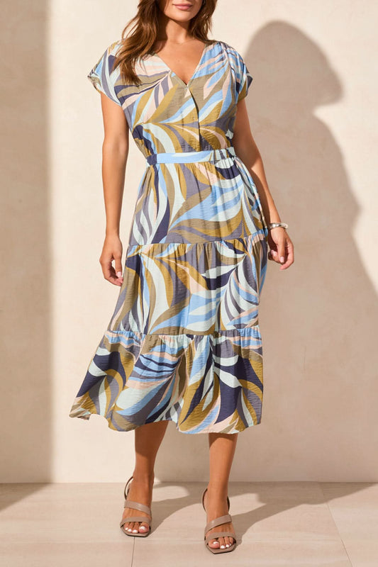 A woman wears the Tribal SHORT SLEEVE V NECK TIERED DRESS with blue, beige, and brown swirls, paired with tan heeled sandals, standing against a light-colored wall.