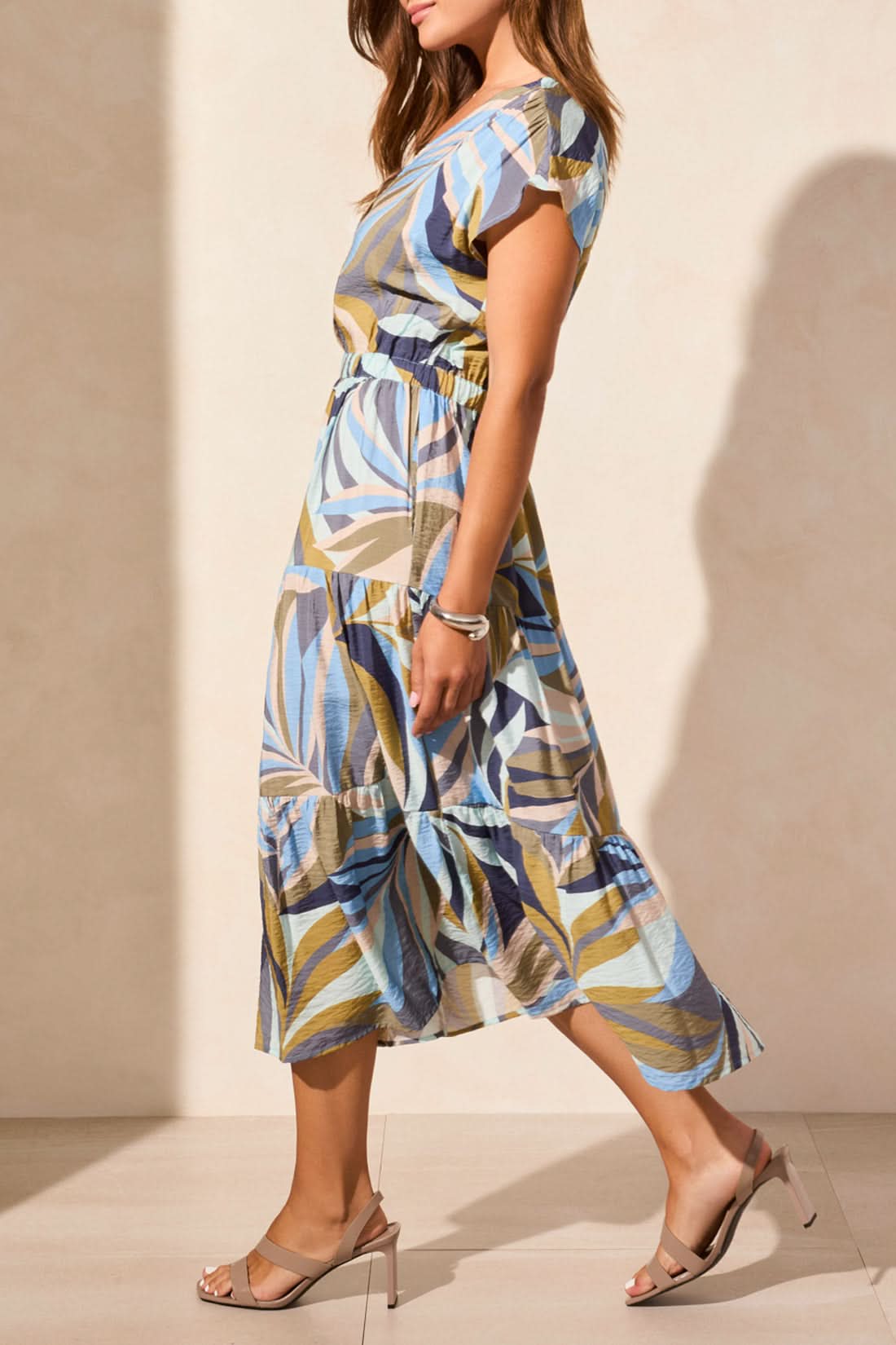 A woman wears the Tribal SHORT SLEEVE V NECK TIERED DRESS with blue, beige, and brown swirls, paired with tan heeled sandals, standing against a light-colored wall.